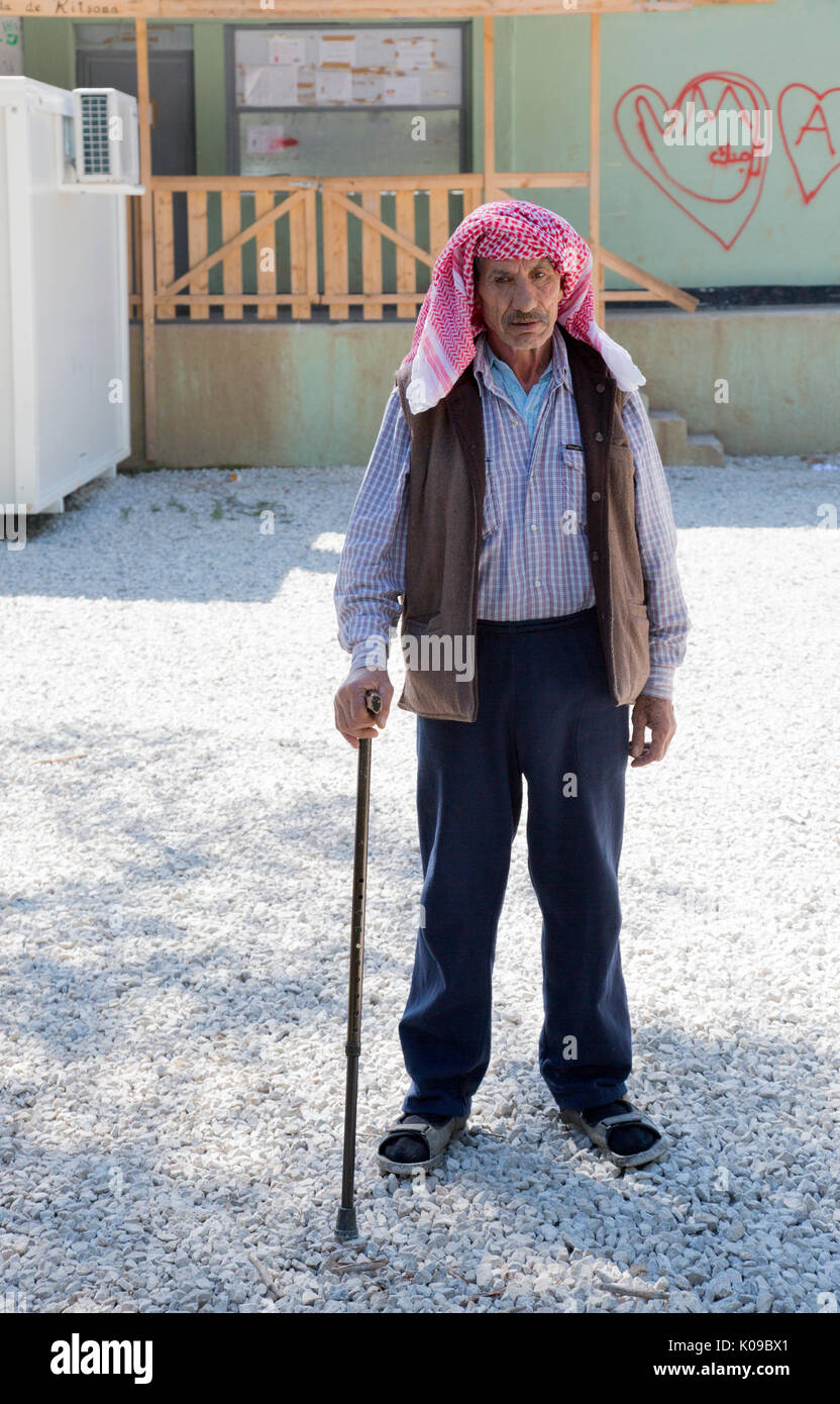 En Ritsona campo de refugiados sirios. Un viejo hombre sirio respaldado por un bastón se sitúa delante de un edificio con graffiti. Foto de stock