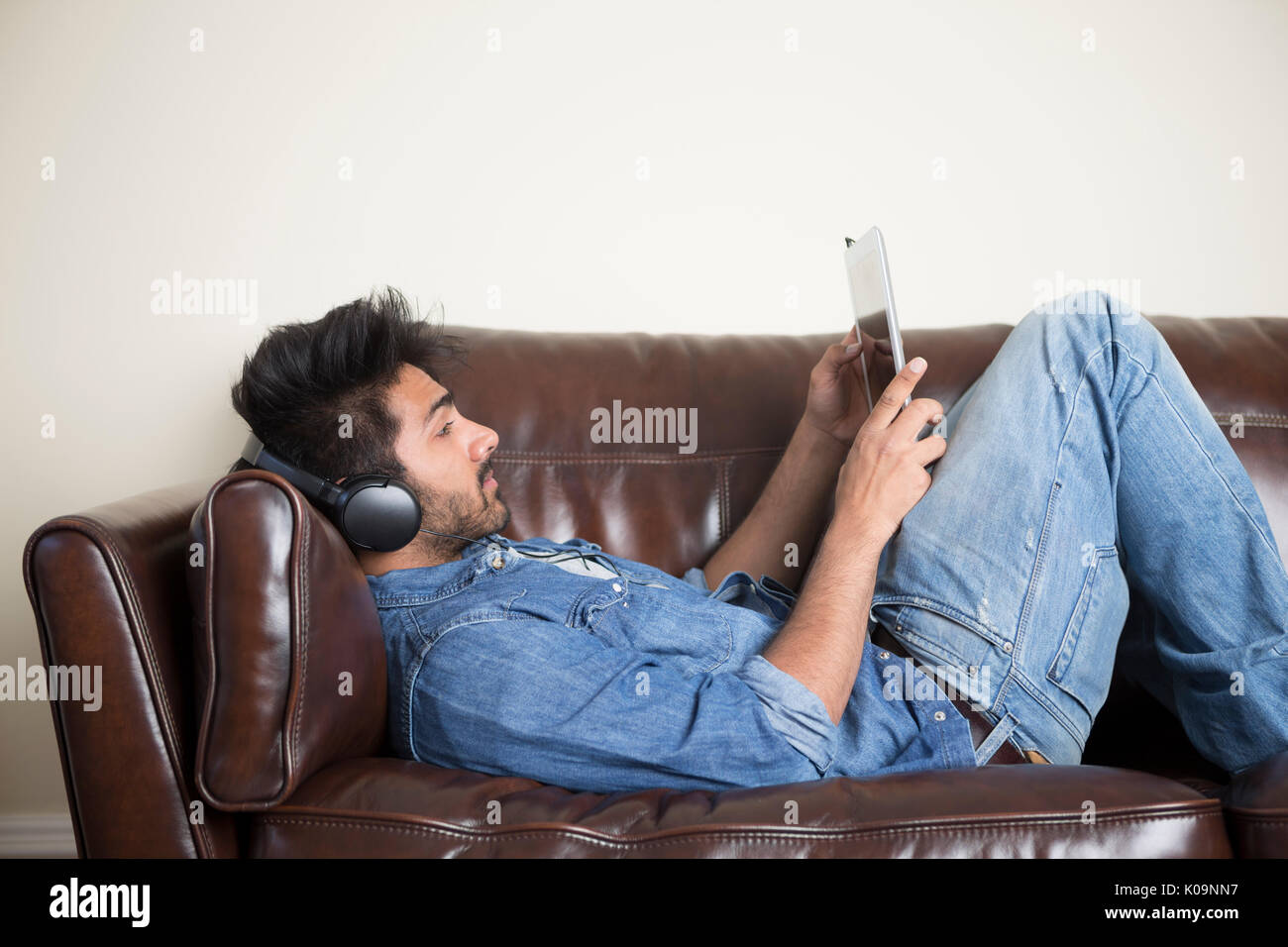 Hombre asiático mediante tableta digital en casa en el sofá usando auriculares. Foto de stock