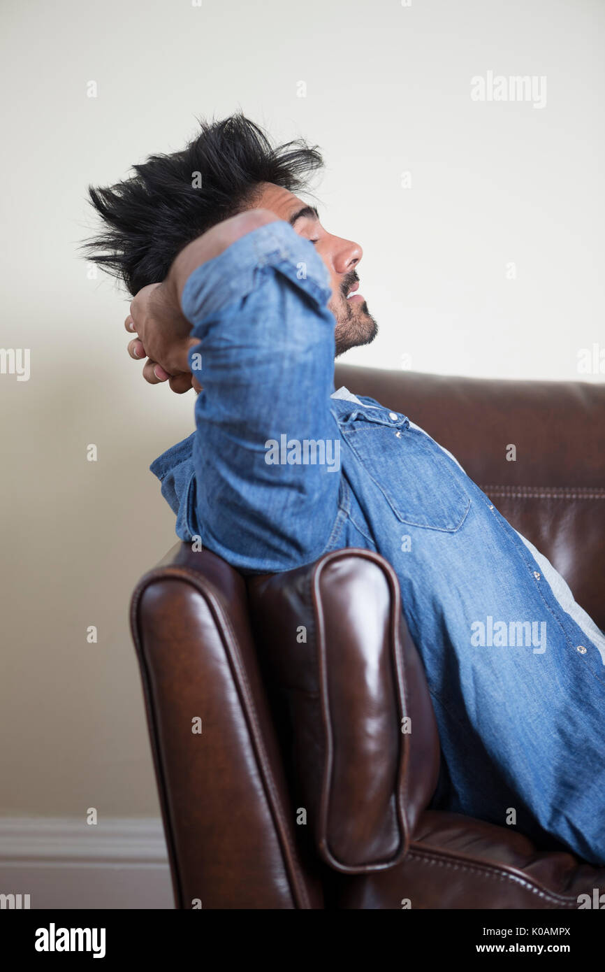 Vista lateral de un cansado o desgraciado hombre asiático sentado en el sofá sosteniendo su cabeza. Foto de stock