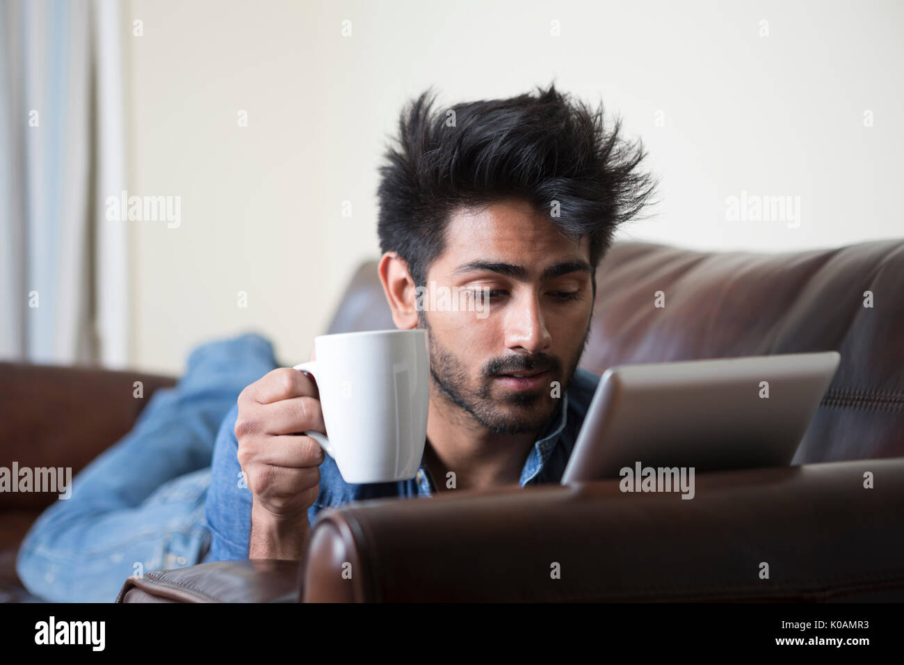 Feliz hombre asiático en casa con una taza de café caliente y usar tableta digital en casa en el sofá. Foto de stock
