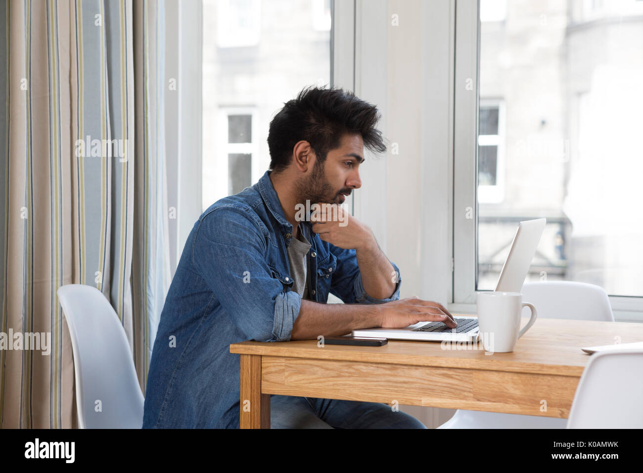 Retrato de un hombre de origen indio sentado a la mesa en casa trabajando en un ordenador portátil. Vista lateral. Foto de stock
