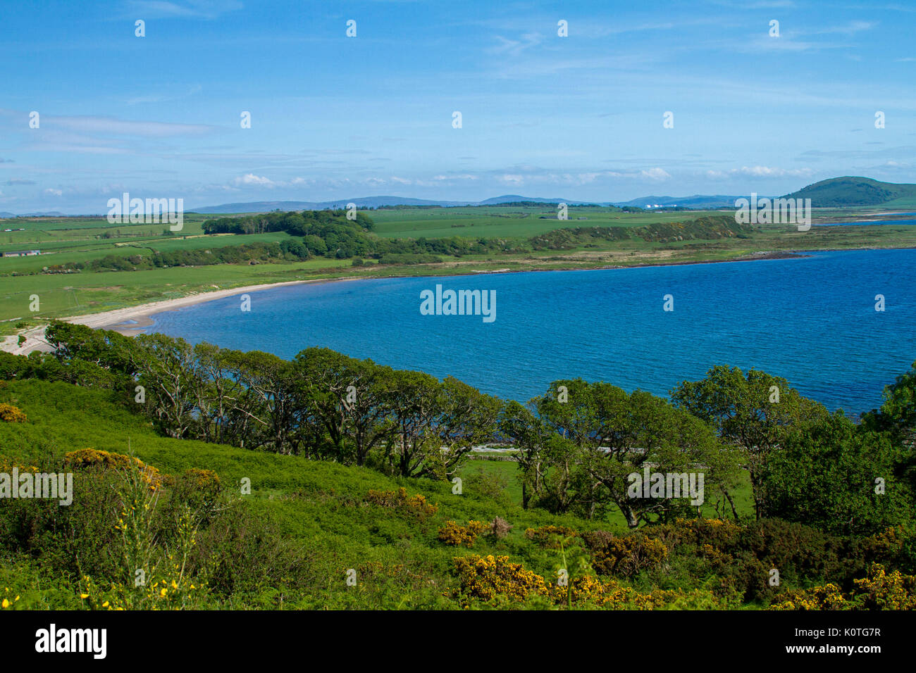El paisaje costero con vista de Scalpsie playa Esmeralda, campos, árboles bajos en la isla de Bute, con montañas de Escocia continental más allá del océano azul Foto de stock