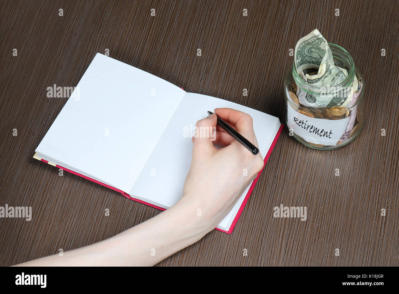 Frasco de vidrio con fondo de retiro lleno de dinero y mano femenina escrito en el bloc de notas Foto de stock