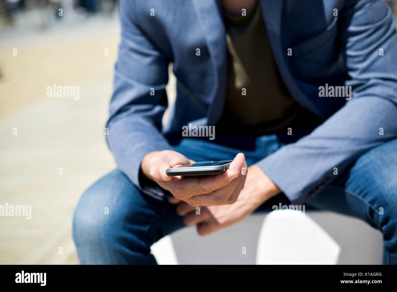 Primer plano de un joven hombre caucásico, vistiendo un elegante casual look,  un par de jeans y una elegante chaqueta azul, utilizando un smartphone  junto a la playa Fotografía de stock -