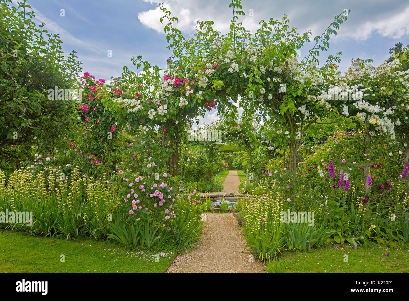 Extensa e impresionante jardín inglés con estanque circular, bordes, y el camino que lleva a través de la arcada con rosas trepadoras en jardines Rousham Foto de stock
