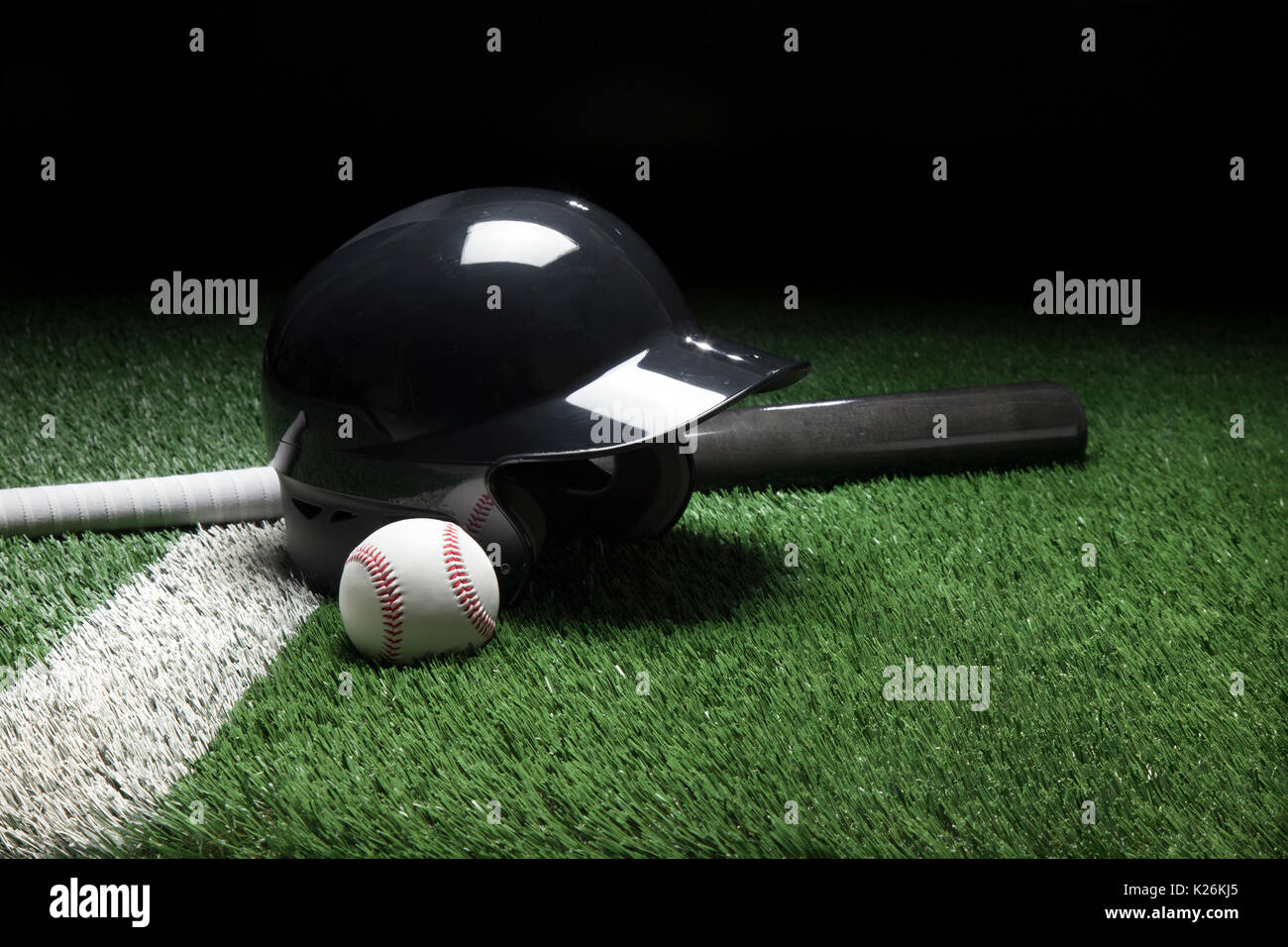 Un casco de bateo de béisbol bate y pelota en un campo con una franja blanca y un fondo oscuro Foto de stock