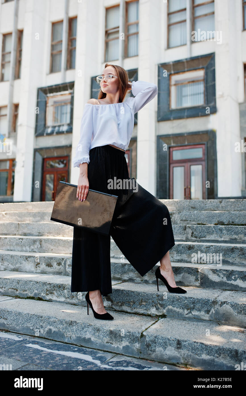 Retrato de una joven mujer en impecable blusa blanca, un pantalón negro ancho y classic high heels posando en las escaleras negro para portátil en la mano con un Fotografía de