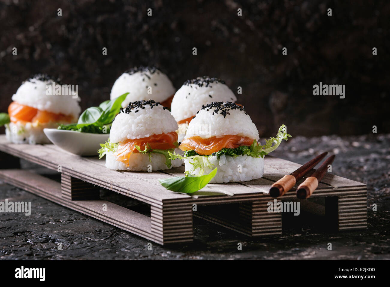Mini hamburguesas de sushi de arroz con salmón ahumado, ensalada y salsas  verdes, sésamo negro servido en bandeja el palet de madera de color marrón  oscuro con palillos sobre hormigón b Fotografía