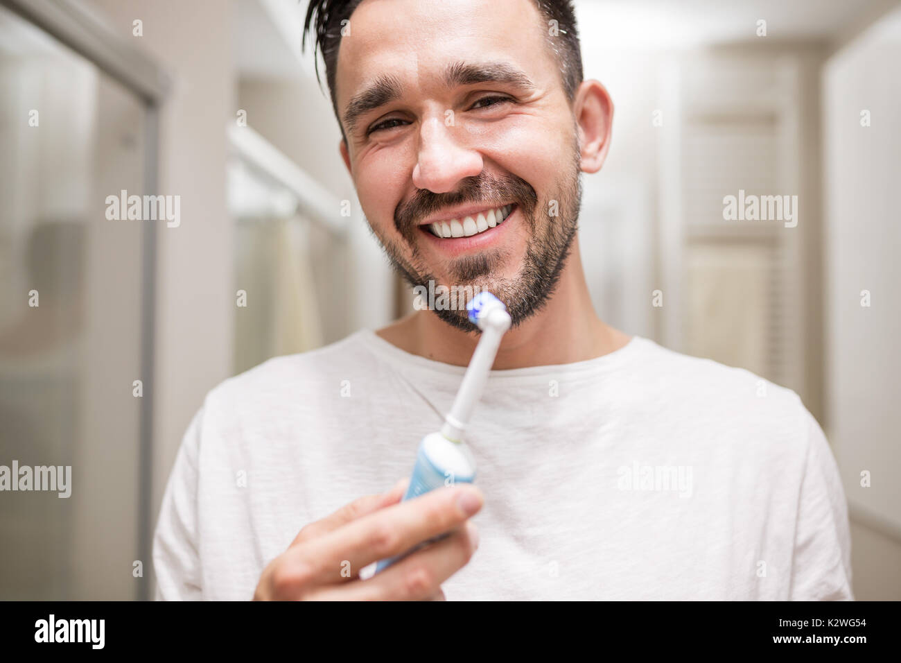 Guapo latino Hombre sujetando un cepillo dental eléctrico y sonriente. Él tiene el pelo negro, barba y vestidos de blanco pijama. Baño interior está en la b Foto de stock