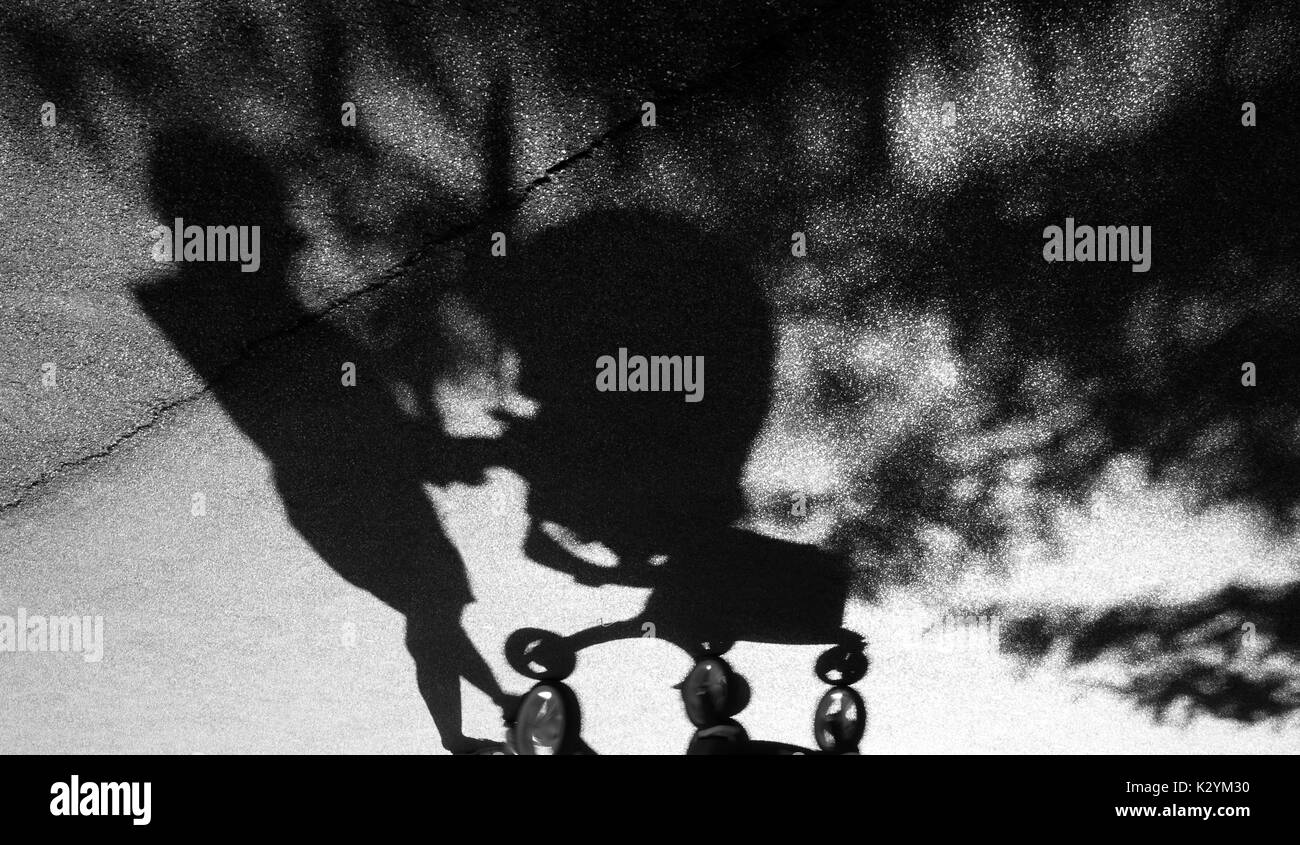 Sombra borrosa de una mujer con depresión posparto empujando un carrito de bebé en el asfalto agrietado suburban Park Road, en blanco y negro Foto de stock