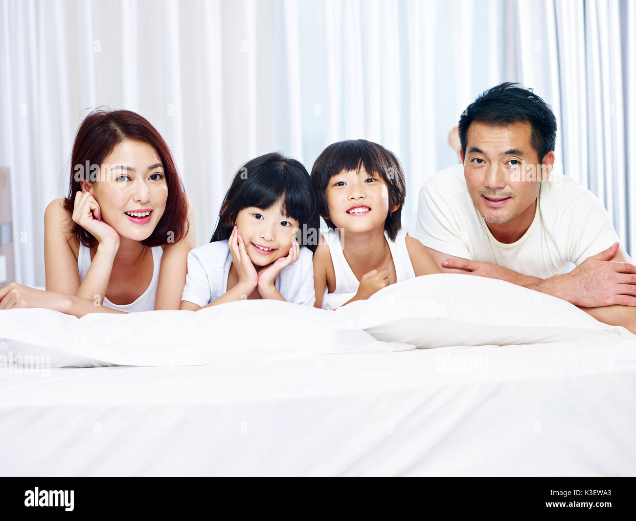 Feliz familia con dos niños asiáticos tumbado en la cama en la parte delantera mirando a la cámara sonriendo. Foto de stock