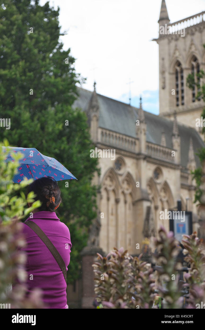 Tourist con paraguas azul se encuentra en el jardín, cerca de Trinity College en cambridge mientras llueve suavemente Foto de stock