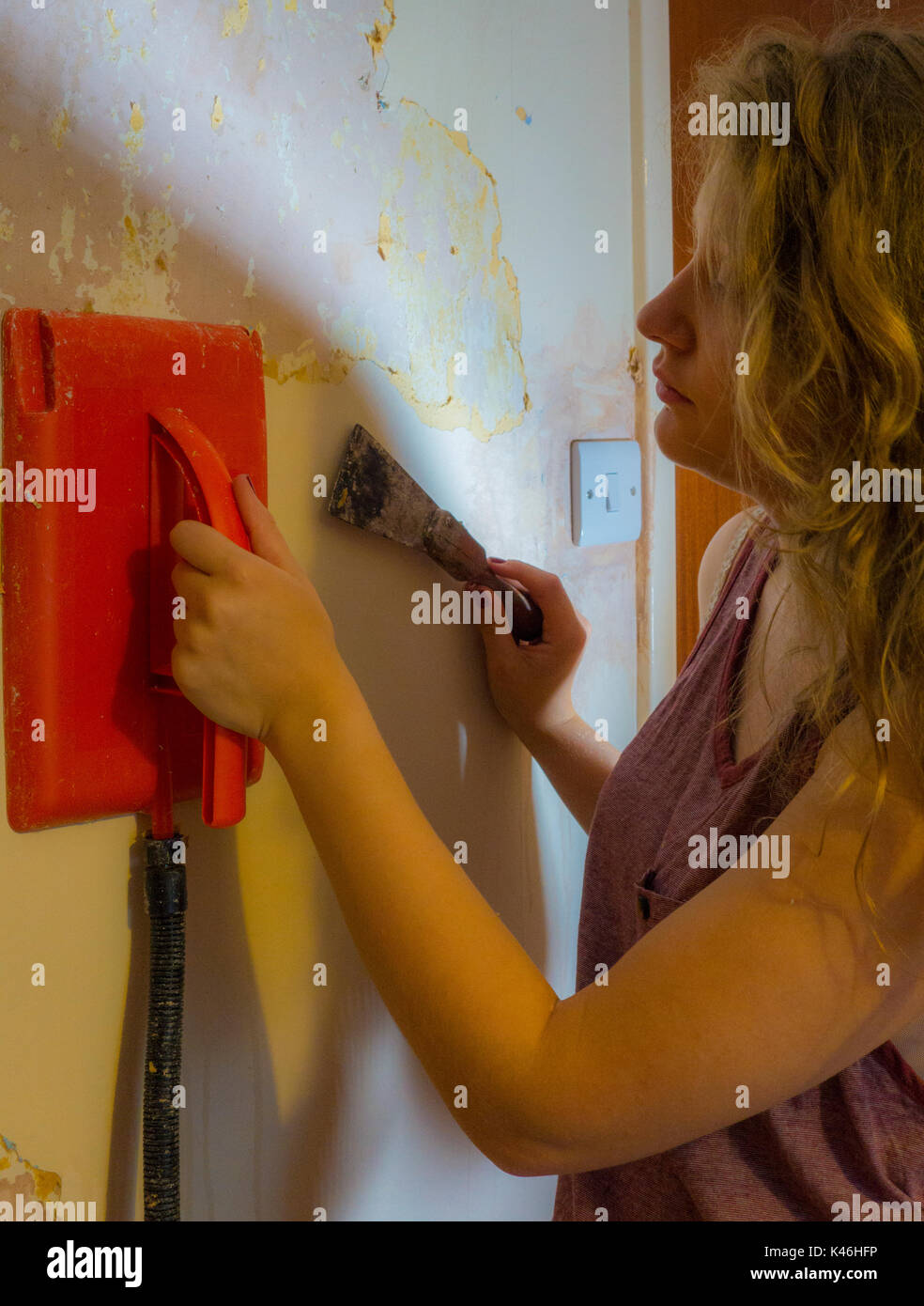 / Decoración redecorar: un bonito joven mujer sosteniendo un vaporizador eléctrico contra una pared, mientras que el papel tapiz antiguo de raspado de una habitación en una casa. Foto de stock