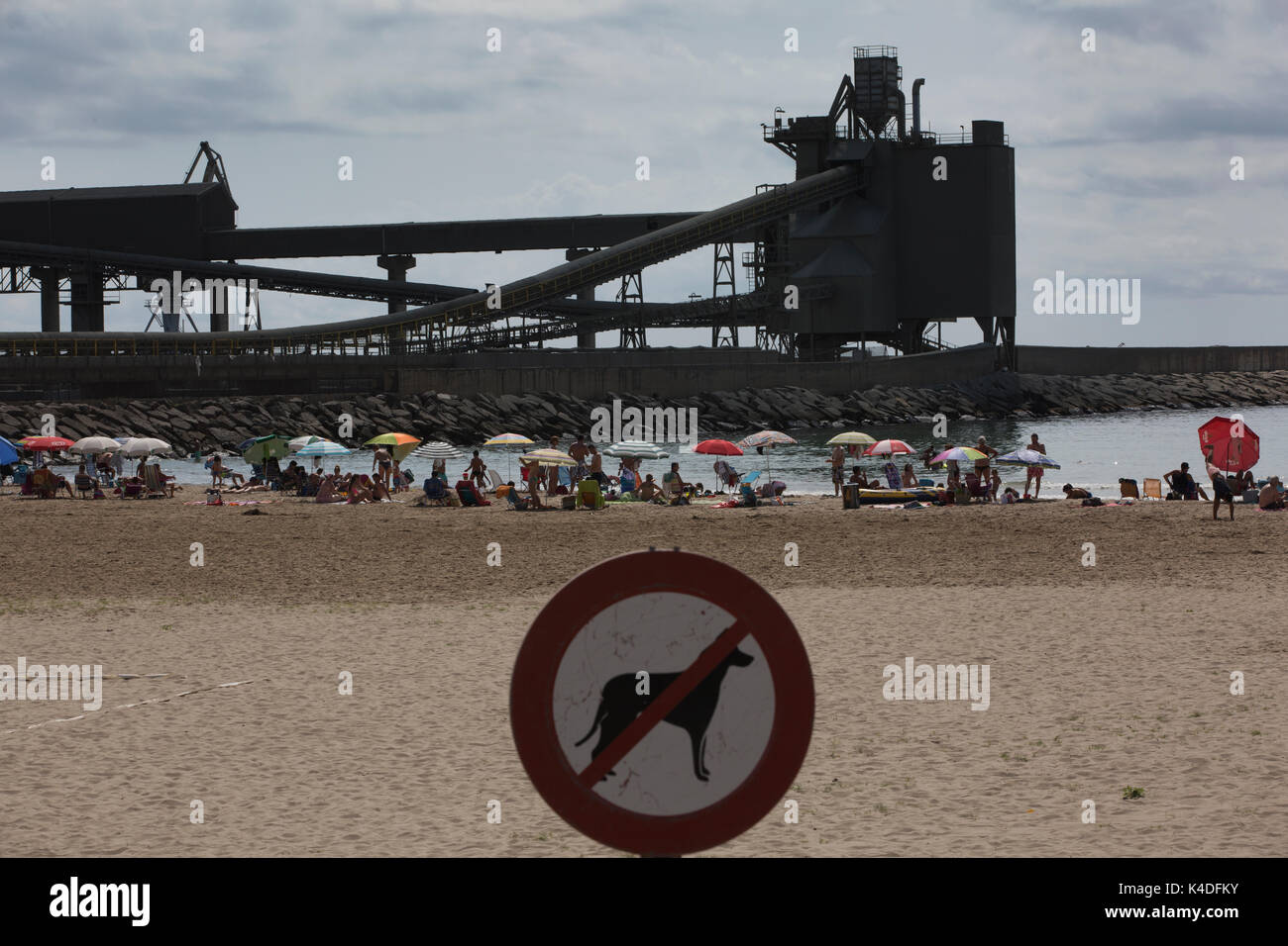 Los turistas disfrutan del verano a lo largo de la playa del Puerto de Alcanar, con vistas a los edificios industriales de la planta cementera Cemex, Alacanar, Tarragona, España Foto de stock