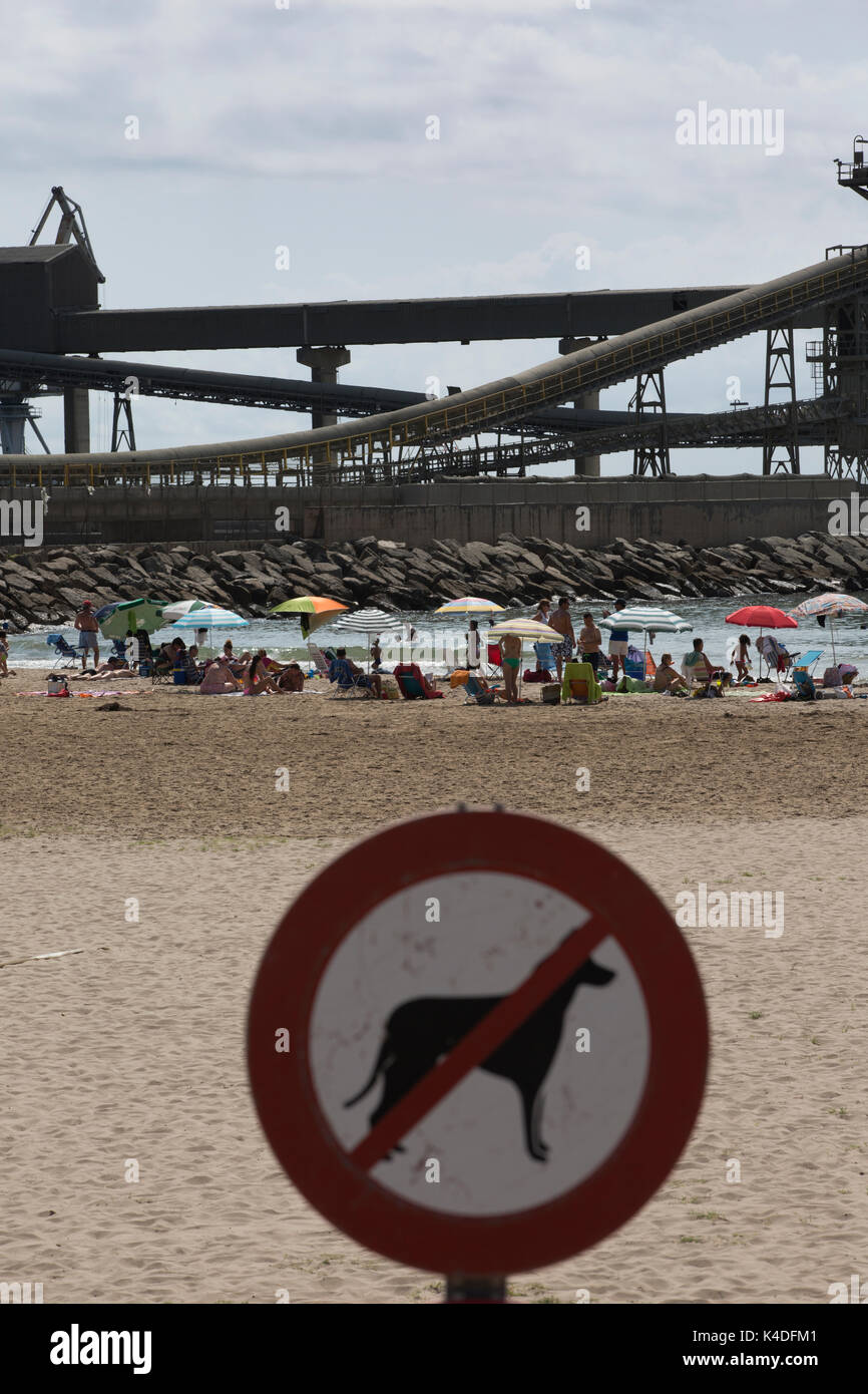Los turistas disfrutan del verano a lo largo de la playa del Puerto de Alcanar, con vistas a los edificios industriales de la planta cementera Cemex, Alacanar, Tarragona, España Foto de stock