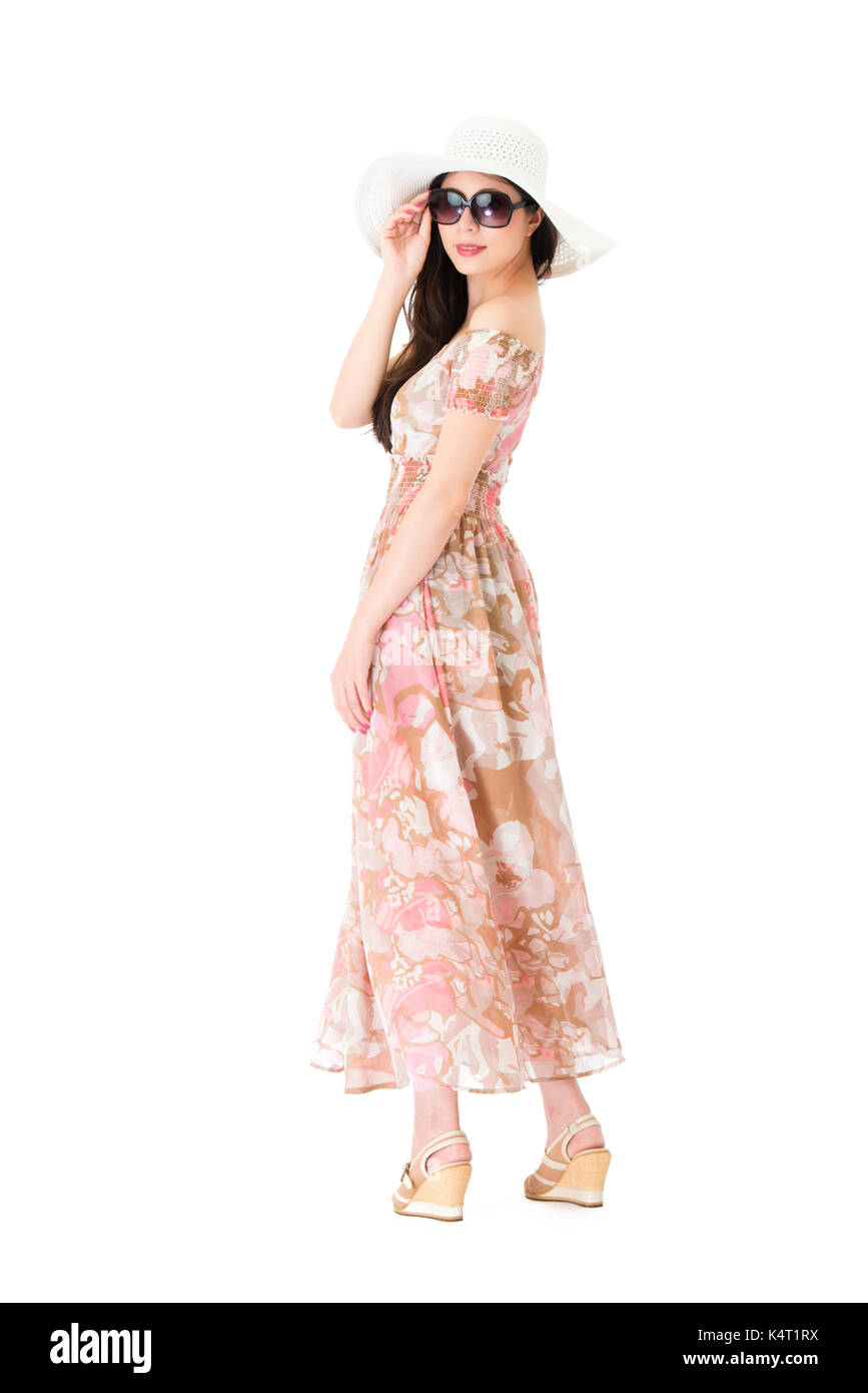 Slim hermosa dama vistiendo ropa de verano vestido con sombrero y girando  el cuerpo mantiene gafas de sol mirando a la cámara de pie sobre la pared  blanca de fondo Fotografía de