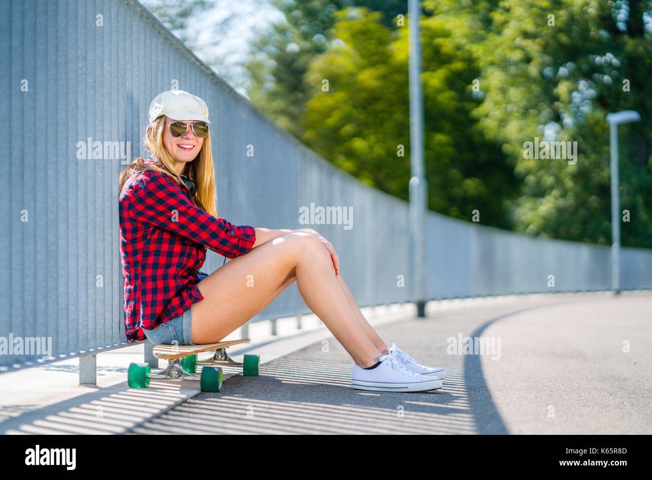 Mujer de 22 años de edad, sentada sobre longboard, se inclina por la calle rejas y relaja Foto de stock