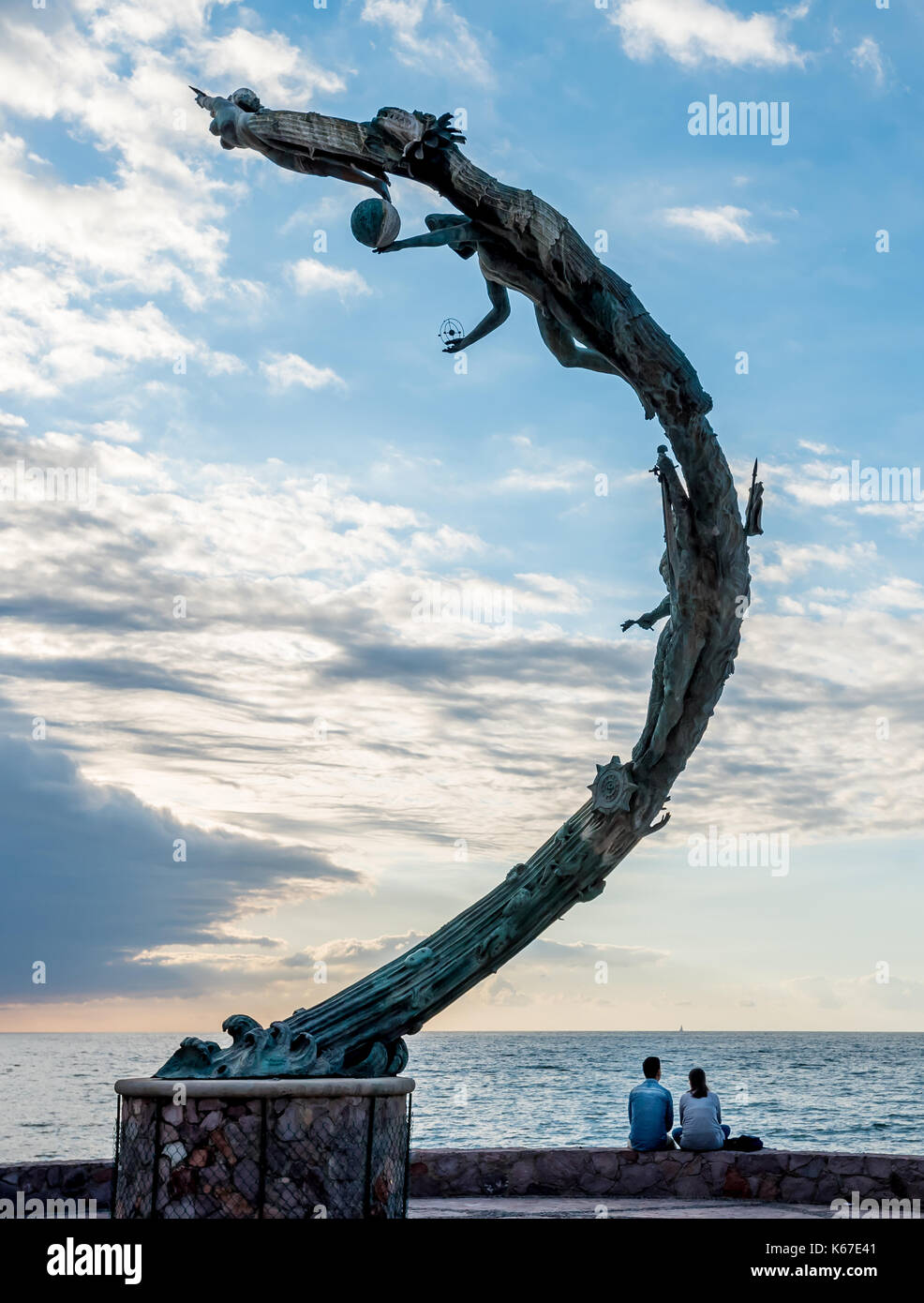 Malecón, Puerto Vallarta, una pareja viendo una puesta de sol sobre la Bahía de Banderas, con el Milenio (milenios) escultura se arquean sobre ellos en las nubes Foto de stock