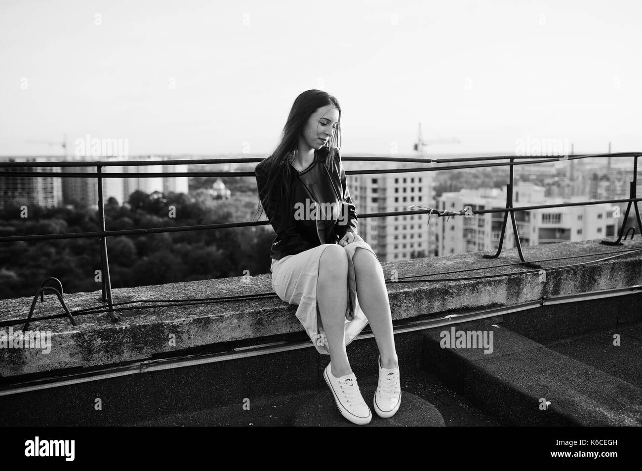 Retrato de una bella muchacha de vestido azul, chaqueta de cuero negro y blanco sneakers sentado en el techo con una hermosa vista de la ciudad. blanco y negro p Foto de stock