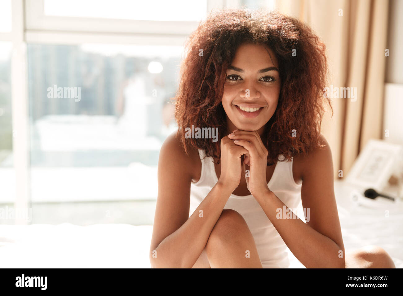 Retrato de mujer africana con el cabello rizado vistiendo pijamas y sonriendo a la cámara en casa Foto de stock