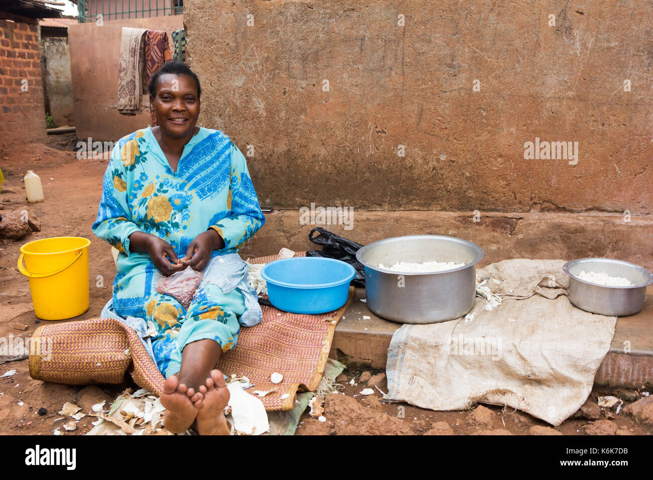 Un negro de mediana edad mujer sentada ugandeses y preparar la comida para cocinar en el frente de su casa. Foto de stock