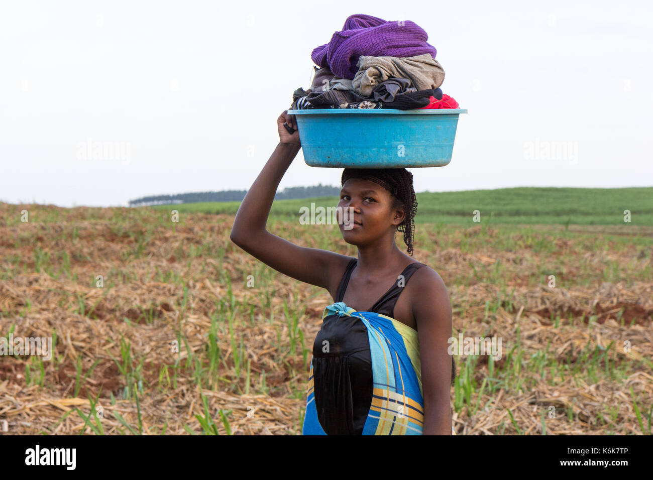 Una joven africana llevando un lavabo lleno de ropa en su cabeza a través de un campo. Foto de stock