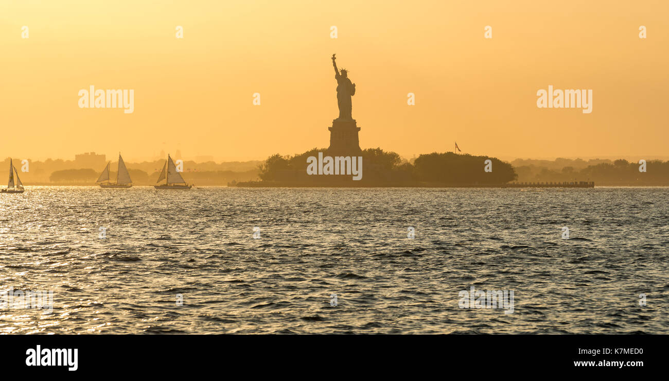 La estatua de la libertad y de la isla de La Libertad a través del puerto de Nueva York con veleros en verano la luz del atardecer. La Ciudad de Nueva York Foto de stock