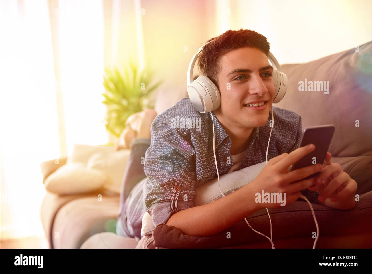 Teen escuchando música con los auriculares del móvil está acostado boca abajo sobre la cama luces multicolores Foto de stock