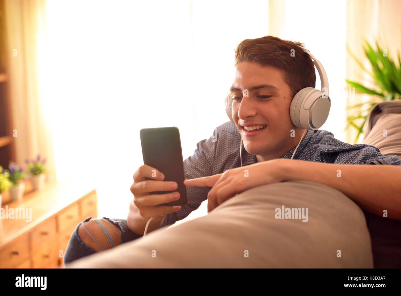 Adolescente ver contenido multimedia en un teléfono móvil con audífonos, apoyándose en la parte trasera de un sofá en casa Foto de stock