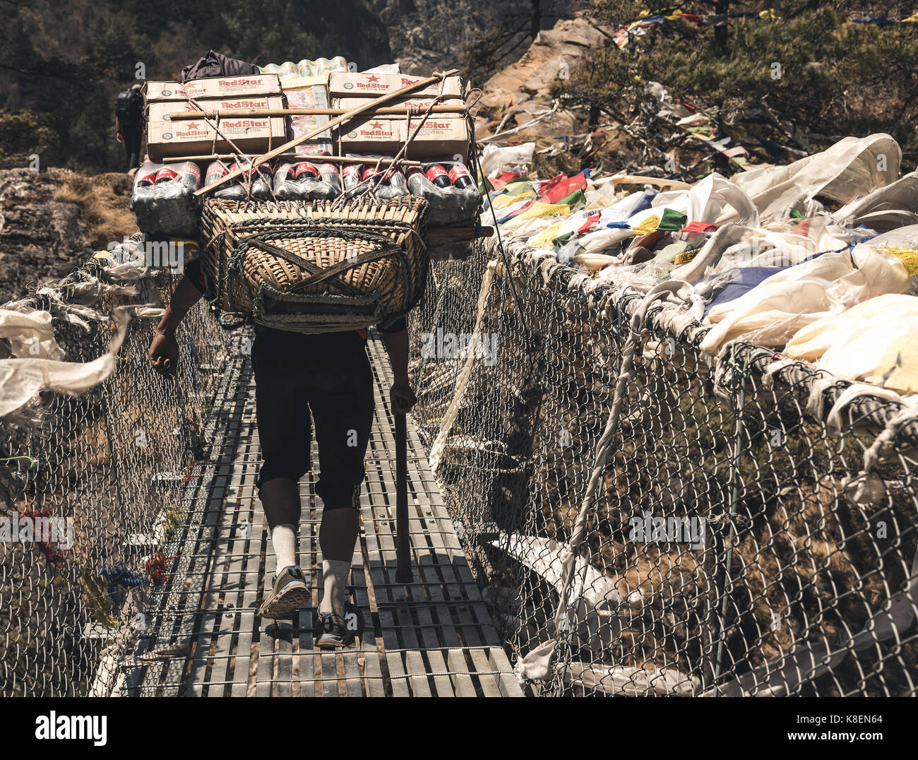 Hombre caminando a lo largo de puente colgante que transporten grandes paquetes de suministros en su espalda. Foto de stock