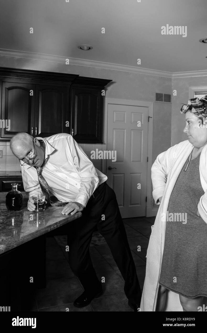 Borracho inclinada contra el contador en cocina con enojado esposa en albornoz y rizadores en blanco y negro. Foto de stock