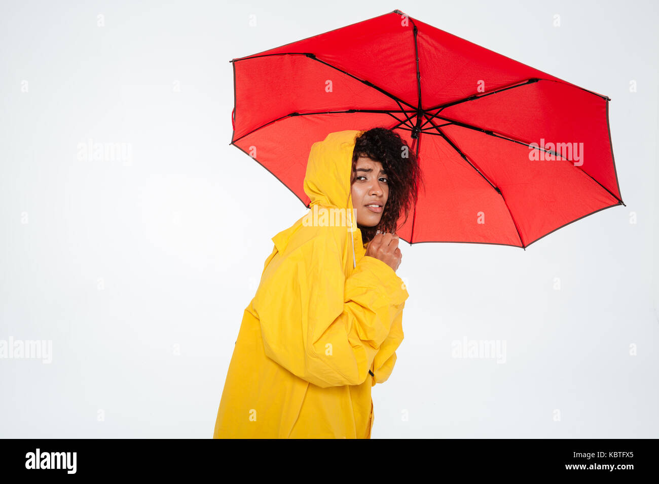 Joven africana en impermeable escondiéndose detrás de paraguas y mirando la cámara sobre fondo blanco. Foto de stock