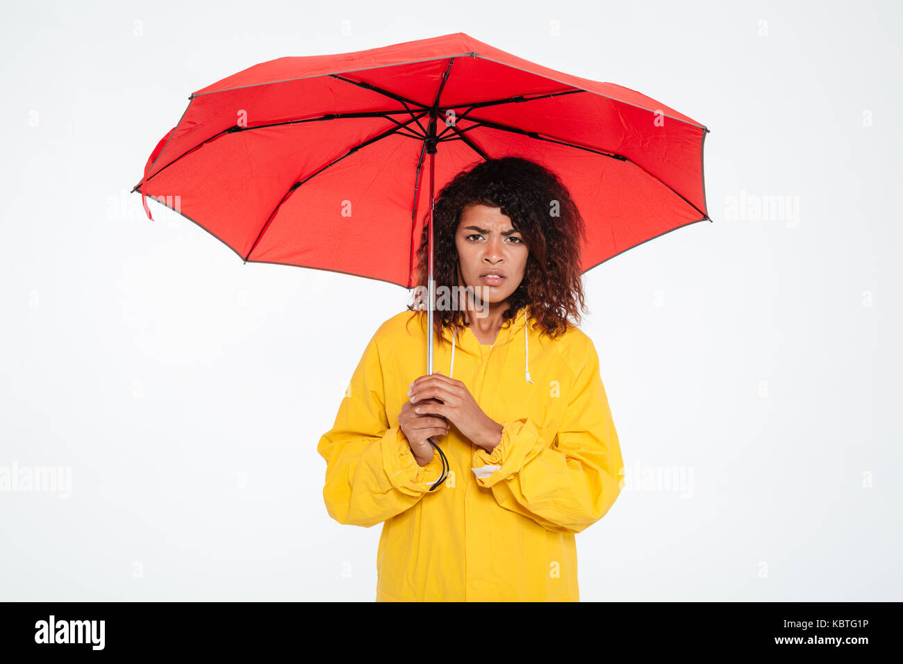 Confundirse mujer africana en impermeable oculto bajo el paraguas y mirando la cámara sobre fondo blanco. Foto de stock