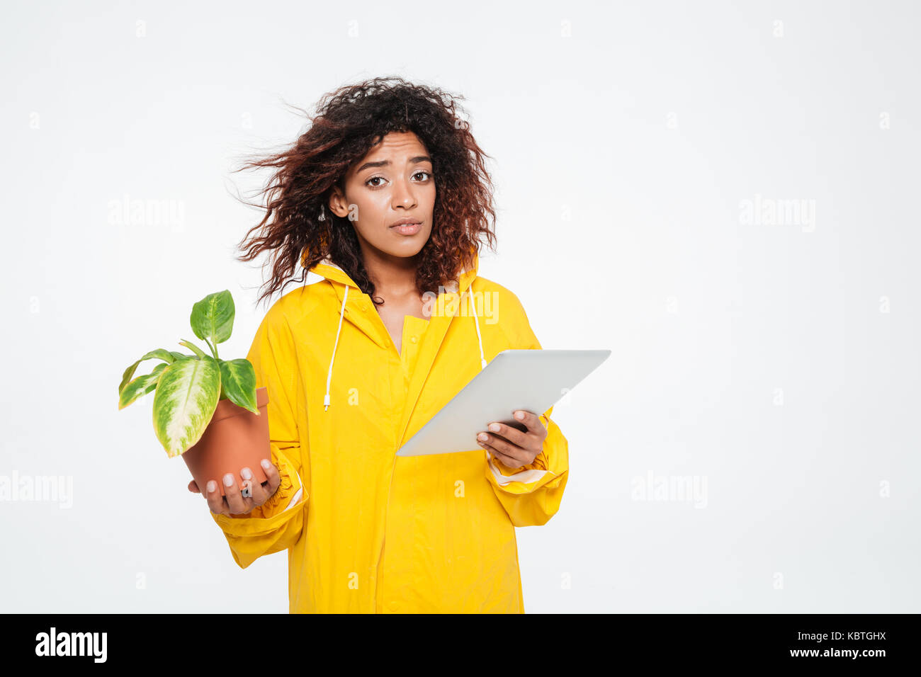 Joven africana en impermeable sosteniendo la planta y mirando la cámara sobre fondo blanco. Foto de stock