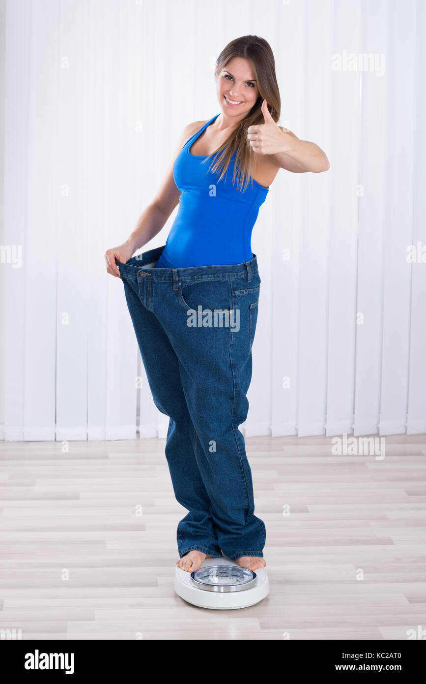 Mujer de slim jeans sobredimensionado de pie sobre la máquina de pesaje en casa Foto de stock