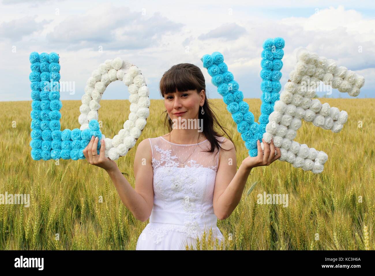 Una chica en un vestido de novia es la celebración de las letras 'amor' de pie contra un campo de trigo y cielo azul. Foto de stock