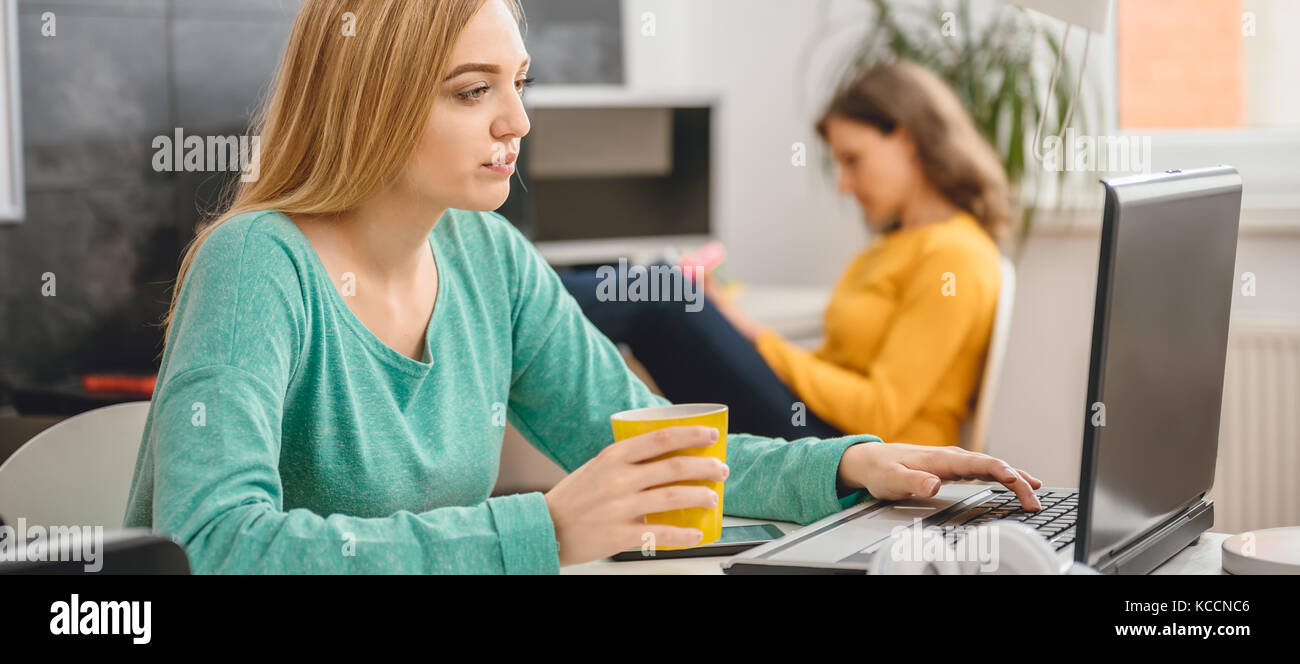 Empresa joven mujer sentada en la mesa, bebiendo café y utilización portátil en el fondo es una mujer por el mostrador tomando notas Foto de stock