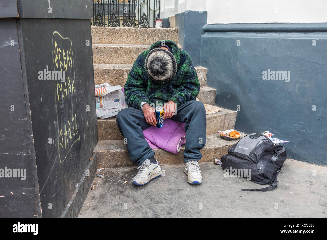 Un hombre sin hogar borracho, sentado en pasos, sosteniendo una lata de cerveza, con una mitad comido comida cerca, con la cabeza hacia abajo y cubierto por un sombrero. Londres, Inglaterra, Reino Unido. Foto de stock