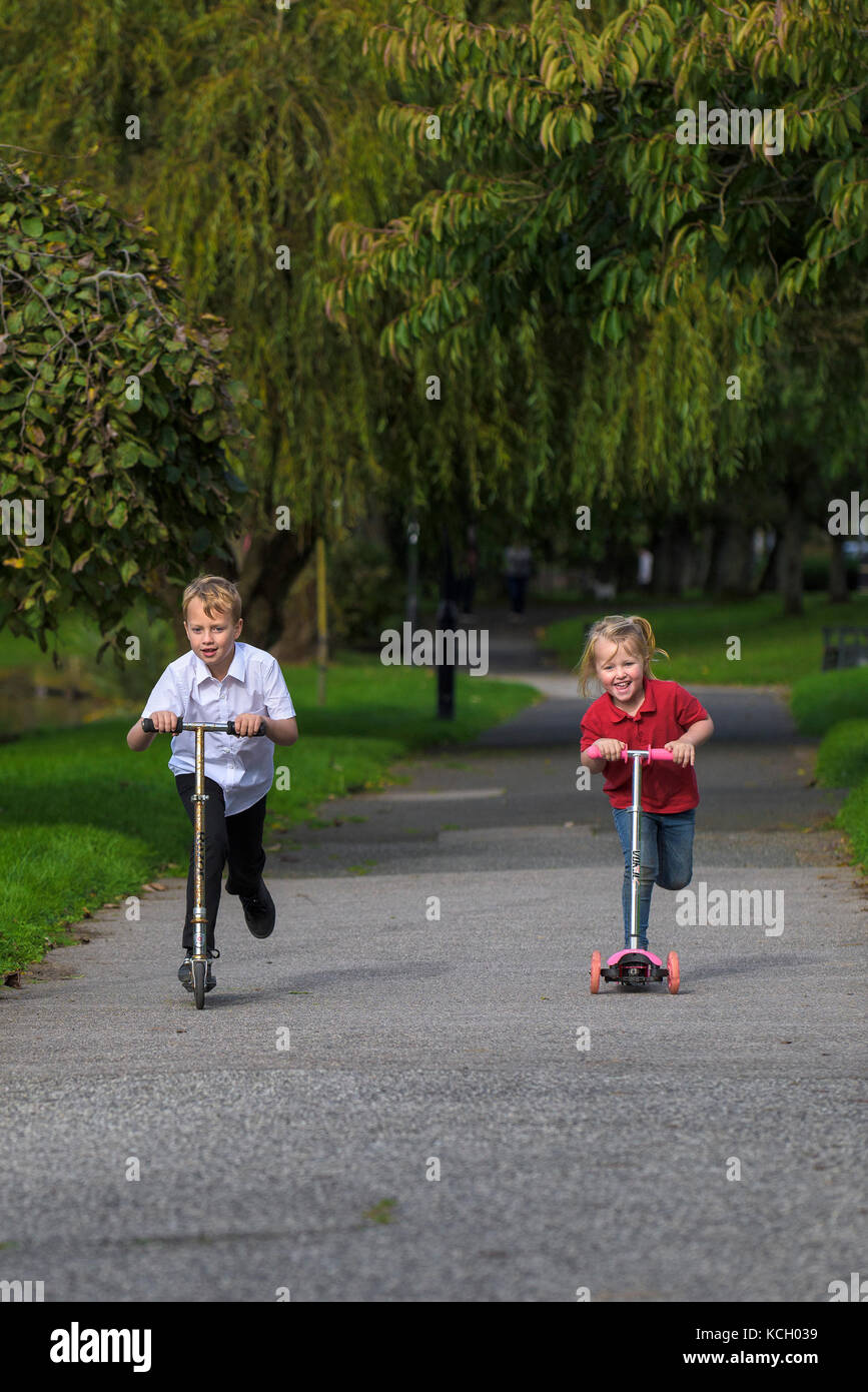 Los niños que viajan sus scooters - un niño pequeño y su hermana joven que se divierten corriendo a lo largo de un camino en sus scooters. Foto de stock
