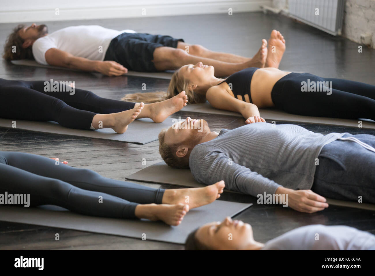Grupo de jóvenes personas practicando yoga lección deportiva con instructor de gimnasio, acostado en el cuerpo muerto, haciendo ejercicio, savasana cadáver plantean, amigos relajante Foto de stock