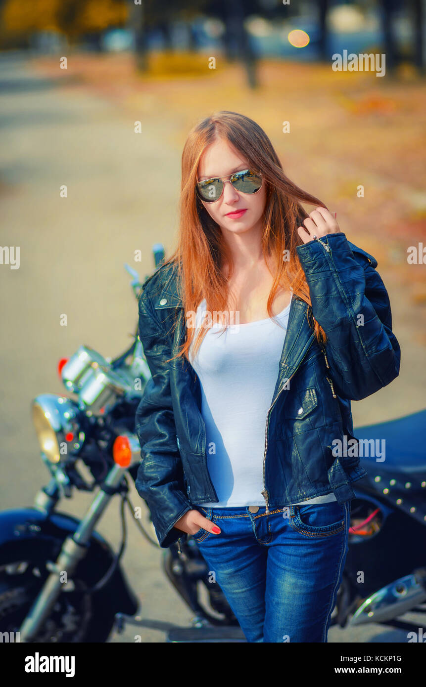 Hermosa joven en una chaqueta de cuero, camisa blanca y pantalones de  mezclilla en una gafas, de pie cerca de la moto en un otoño urbano  superficial de fondo d Fotografía de