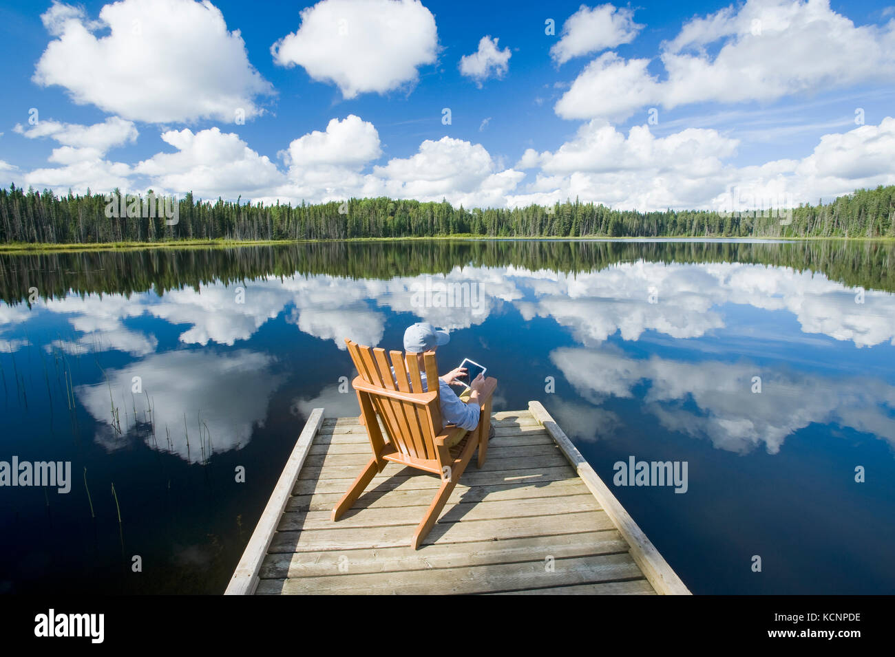 Hombre sentado en una silla en el muelle con un tablet, Two Mile Lake , Pato Parque Provincial de montaña, Manitoba, Canadá Foto de stock