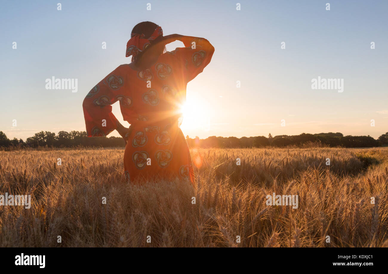 Mujer africana con ropa tradicional de pie, mirando a los ojos, la mano, en el campo de cebada o trigo al atardecer o al amanecer Foto de stock