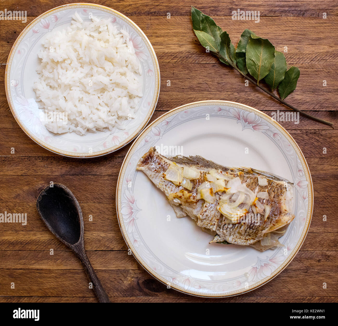 Microondas cocinan los pescados enteros, con ajo y cebolla en rodajas  encima, sobre una placa decorativa de porcelana blanca y un plato de arroz  blanco sobre un fondo de madera ligera Fotografía
