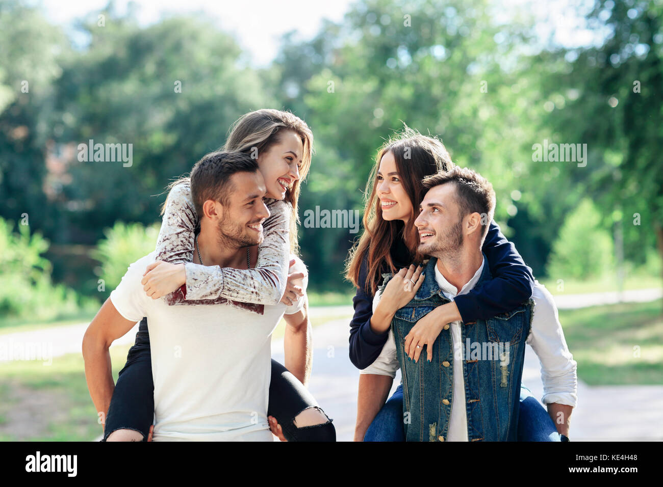 Dos enamorados piggyback divertirse al aire libre. Las niñas sentados en la espalda de chicos sonriendo. mujeres y hombres jóvenes jugando en el parque, y mirando cada int Foto de stock