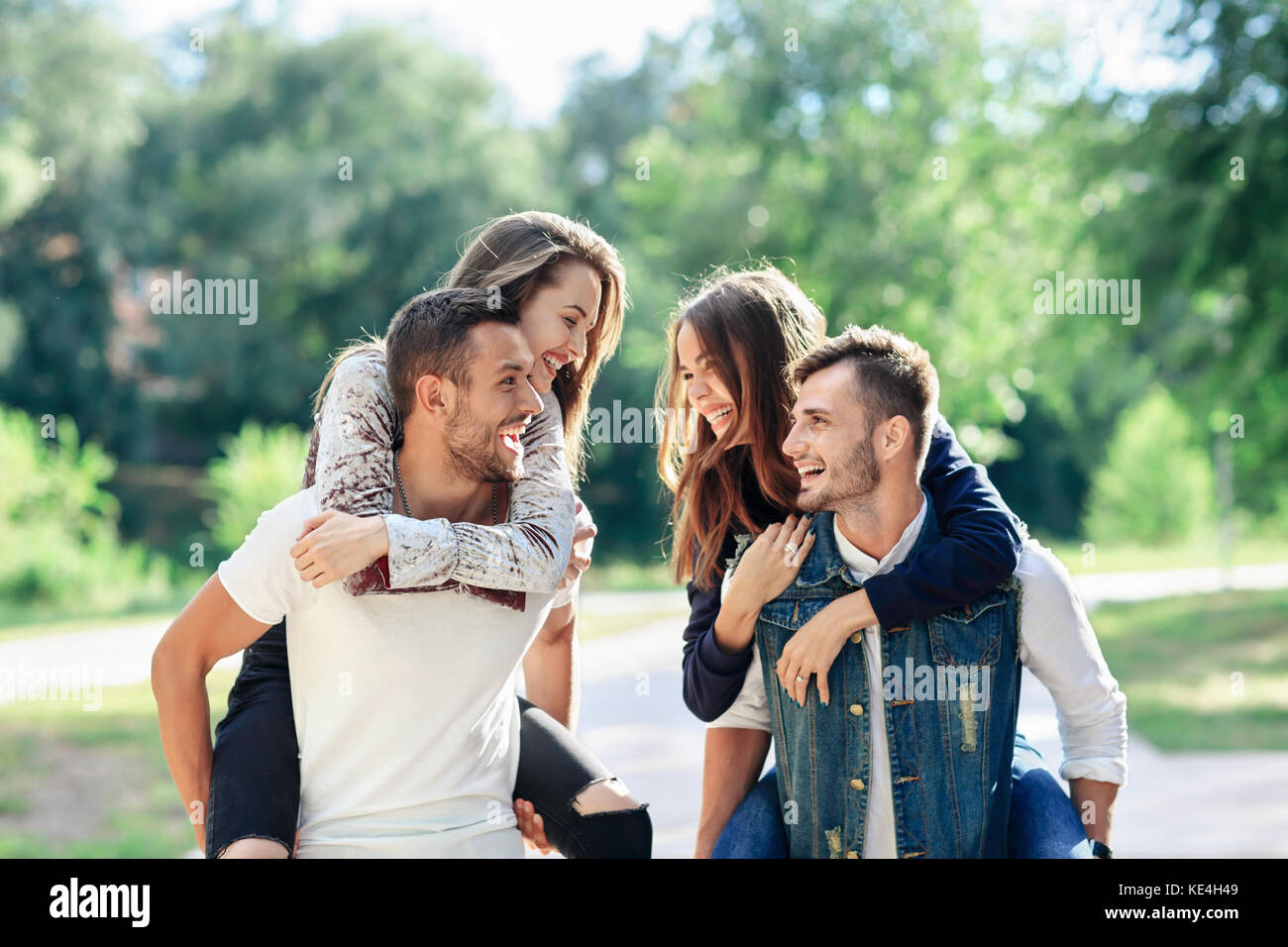 Dos enamorados piggyback divertirse al aire libre. Las niñas sentados en la espalda de chicos sonriendo. mujeres y hombres jóvenes jugando en el parque, y mirando cada int Foto de stock