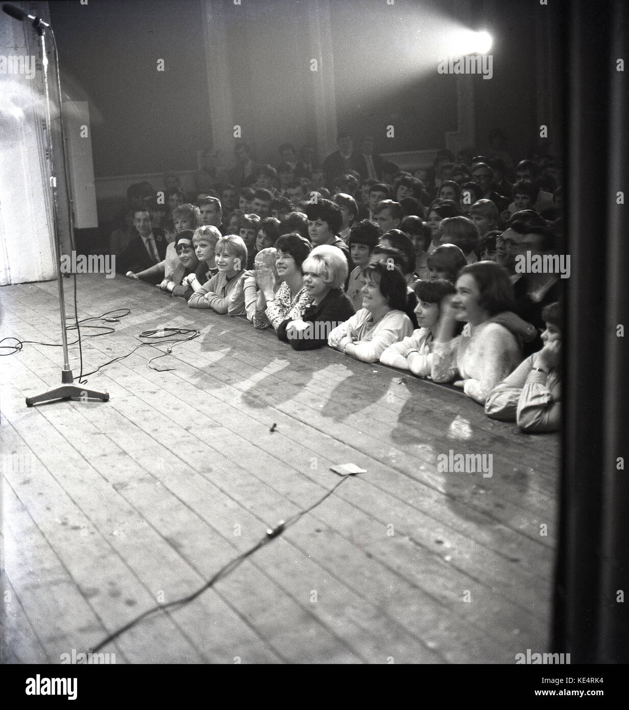 1964, históricos, un micrófono está en la fase de madera en la Bah, Aylesbury, como la audiencia de jóvenes adultos, principalmente mujeres jóvenes, presione contra la parte delantera del escenario en ansiosos anticpation de la noche de ejecución musical, Inglaterra, Reino Unido. Foto de stock