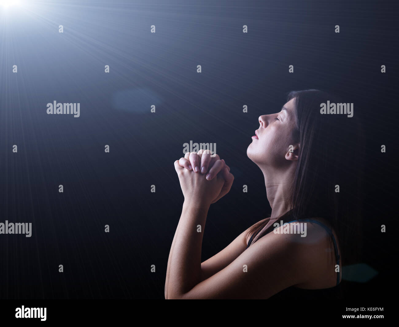 Cerca de una mujer fiel orando bajo una luz celestial o divino, manos plegada en el culto, la cabeza hacia arriba y los ojos cerrados en oración el fervor religioso. Foto de stock