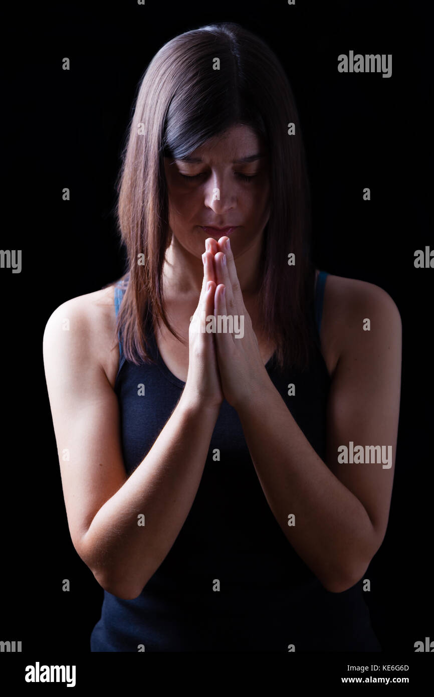 Fiel athletic mujer orando, con las manos plegadas en el culto a Dios, con la cabeza hacia abajo y los ojos cerrados en el fervor religioso, en concepto de fondo de color negro. Foto de stock