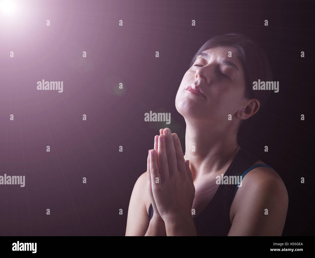Mujer fiel orando bajo una luz celestial o divino y sentir la presencia o ser tocado por Dios. manos plegadas en el culto, cabeza arriba Foto de stock